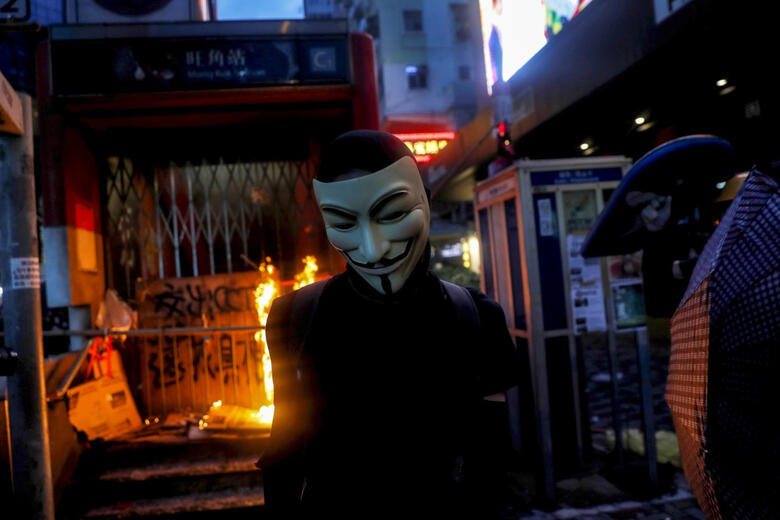 an mtr station burns behind a protester in a guy fawkes mask
